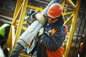 A construction worker carrying a gray pipe at a construction site