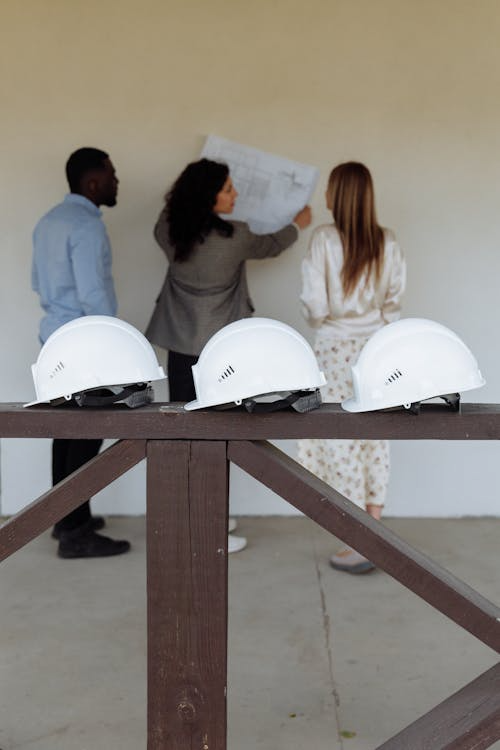 An image of three white helmets in focus while three consultants look at construction blueprints in the background  
