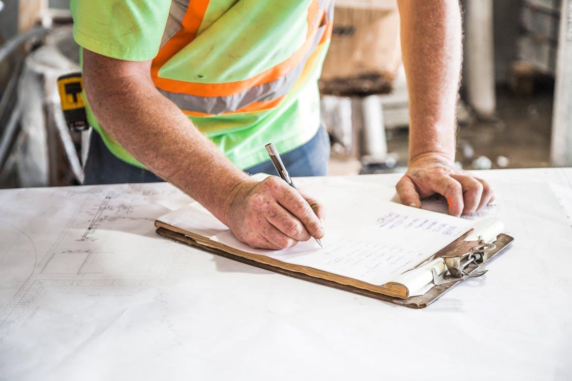 Construction worker reviewing plans and writing notes on a clipboard at a job site.